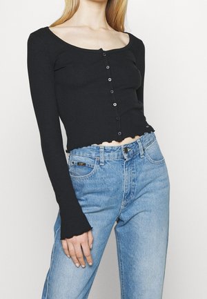 Frau trägt ein eng anliegendes schwarzes langärmeliges, mit Knöpfen versehenes Croptop und eine hoch taillierte hellblaue Jeans vor einfarbigem Hintergrund.