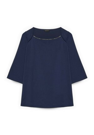 Marineblaue Bluse mit langen Ärmeln und einem Rundhalsausschnitt, der mit einer Reihe kleiner silberner Verzierungen über der Brust versehen ist.