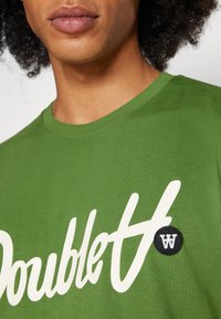 Green cotton t-shirt featuring a white cursive logo and a black circular accent with a white 'W' on the left chest area.