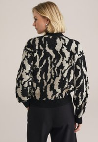 Överdimensionerad stickad tröja i beige och svart abstrakt mönster, med ribbad krage och muddar, visad bakifrån med sänkta axlar.