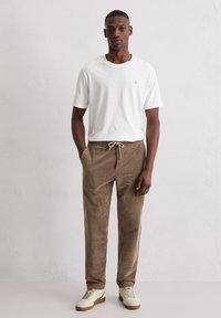 Mann steht mit den Händen in den Taschen, trägt ein weißes kurzärmliges T-Shirt, braune Jogginghosen mit Kordelzug und weiße Turnschuhe vor einer schlichten Wand.