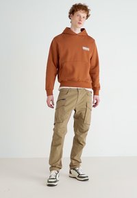 Bruine capuchontrui met kangoeroezak, gecombineerd met tan cargo-broeken met meerdere zakken en donkere accenten. Gemaakt met sneakers.