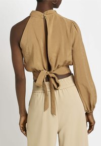 Ljusbrun kort blus med knyt i ryggen, puffiga långa ärmar och hög krage; ihop med beige högmidjade byxor med resår i midjan.