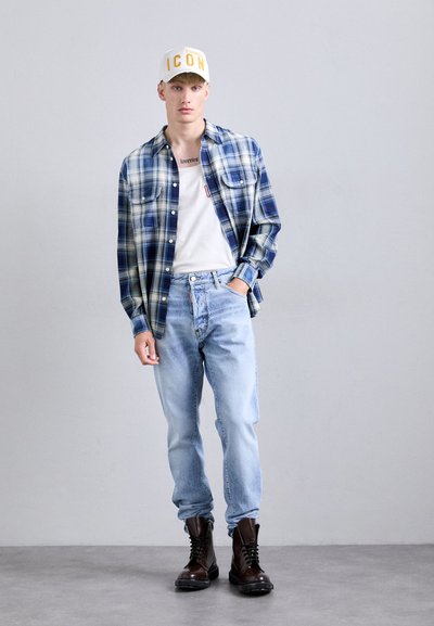 Joven con una gorra blanca que dice "ICON", una camisa de cuadros azul sobre una camiseta blanca, jeans de mezclilla azul claro y botas de cordones marrón oscuro, de pie contra una pared gris.