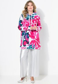 Kápová tunika v živé růžové, tyrkysové a bílé květinové potisku, doplněná o lesklé stříbrné kalhoty s širokými nohavicemi a stříbrné vysoké podpatky.