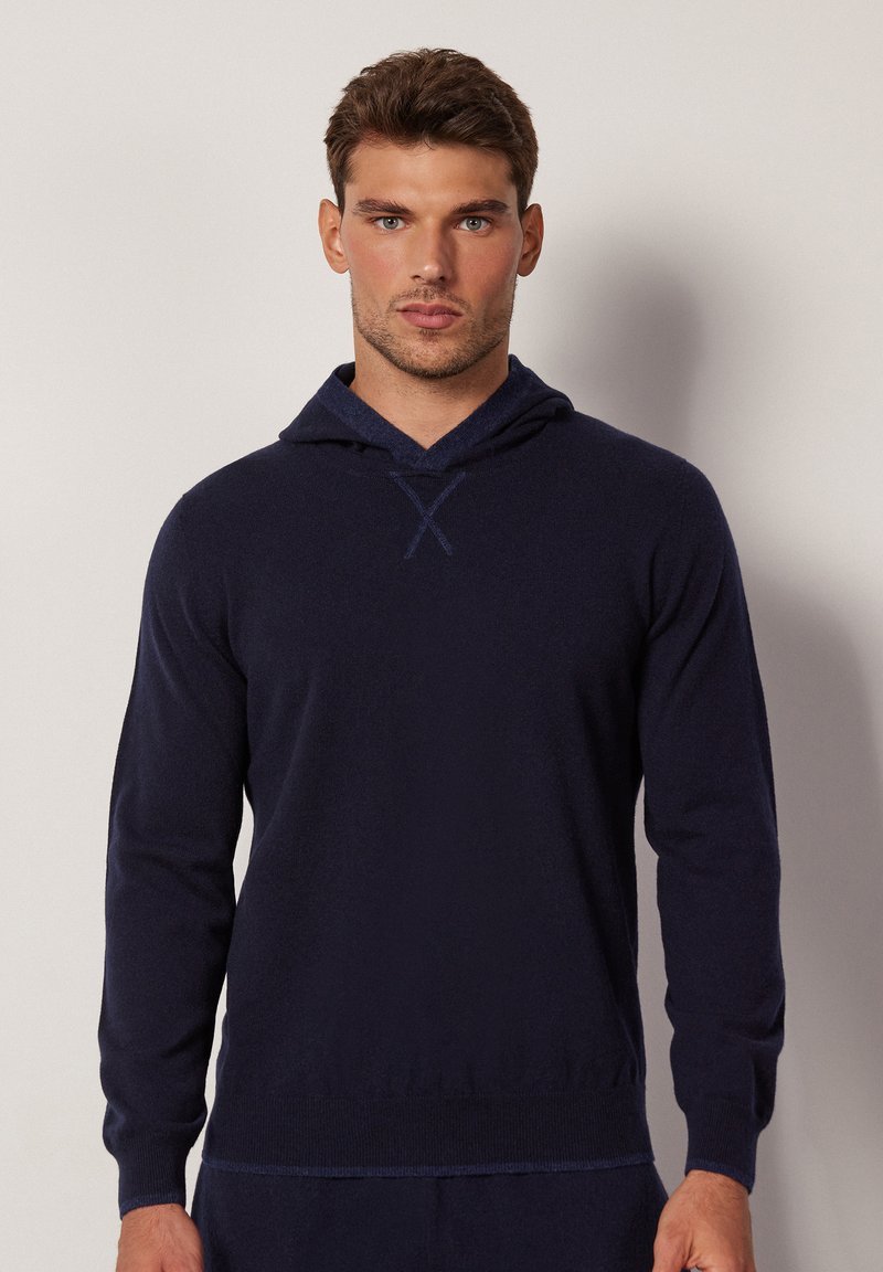 Marineblaue Kapuzenpullover aus weichem Strickmaterial, mit V-Ausschnitt, langen Ärmeln und strukturiertem Saum. Akzente sind ein kontrastierender Kapuzenbesatz.