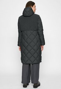 Woman wearing a long black quilted coat with textured upper back and hood, gray wide-legged pants, and black platform shoes, standing facing backward.
