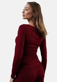 Burgundowa bluzka z długim rękawem, z marszczonym wykończeniem z tyłu, z okrągłym dekoltem i dopasowanym krojem, który tworzy smukłą sylwetkę.