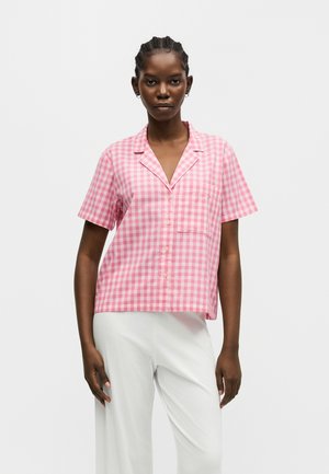 Femeie purtând o cămașă scurtă cu mâneci roz și albe în carouri și pantaloni albi, stând în picioare în fața unui fundal simplu și luminos.