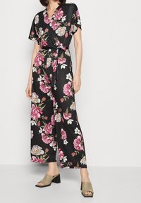 Mono negro con estampado floral, de mangas cortas y cinturón de lazo, que presenta grandes flores rosas y blancas. Combinado con sandalias de tacón cuadrado en color verde oliva.