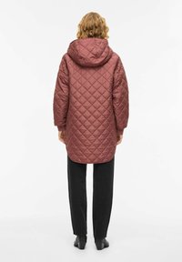 Vattert kåpe med hette i rosarødt, med lang fasong og senkede skulderpartier. Kombinert med svarte bukser og sko med lav hæl.