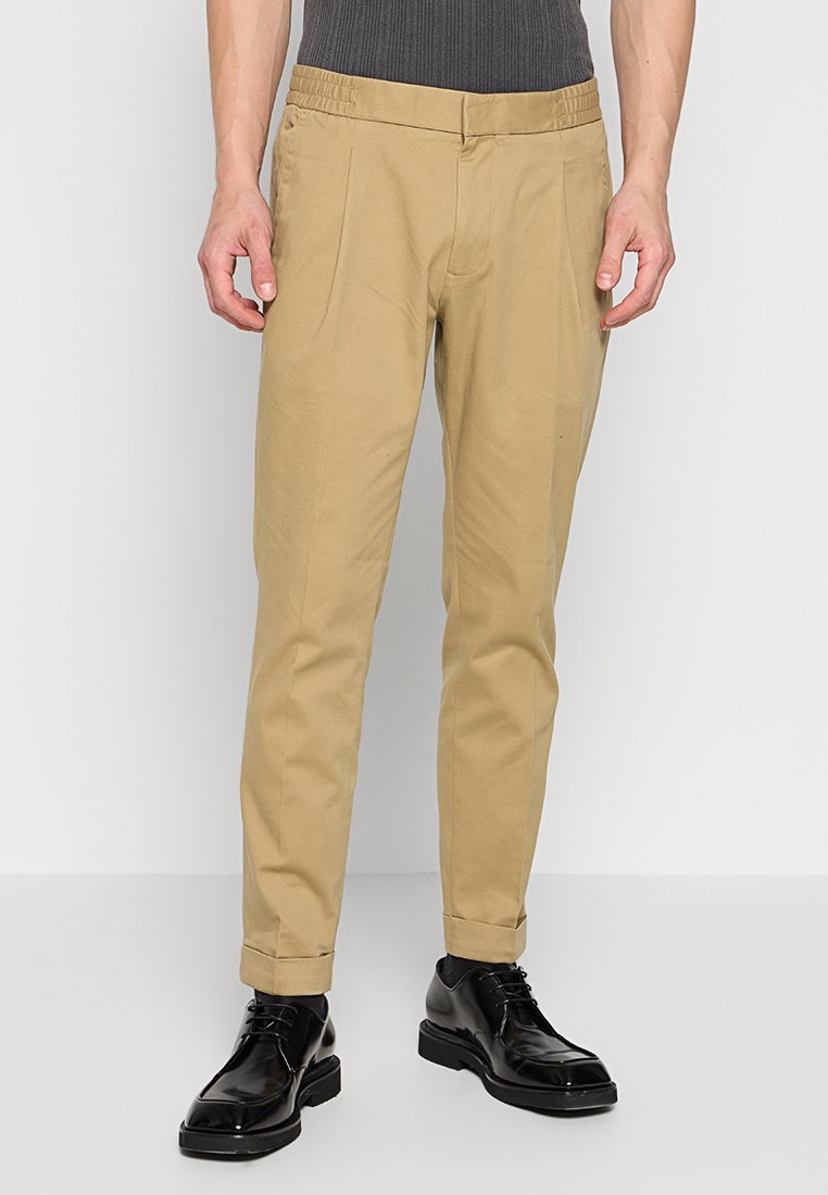Pantalons beiges ajustés avec des plis sur le devant, ourlets retroussés, portés avec des chaussettes noires et des chaussures noires à lacets brillantes.