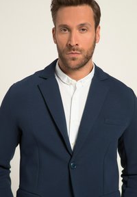 Navyblauer Blazer mit revers, einem Knopfverschluss und einer linken Brusttasche, über einem weißen Kragenhemd getragen.