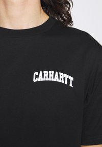 T-shirt en coton noir avec un col rond et des manches courtes, présentant un logo "CARHARTT." blanc en courbe sur le coin supérieur gauche.