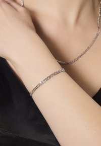 Srebrna bransoletka łańcuszkowa na nagim nadgarstku, o plecionej konstrukcji. Tekstura jest gładka, a kolor błyszczący metaliczny z subtelnymi refleksami.