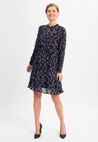 Schwarzes durchscheinendes Kleid mit lila Blumenmuster, langen Ärmeln, Schlüsselloch-Ausschnitt und tailliertem Bund. Kombiniert mit schwarzen High Heels.