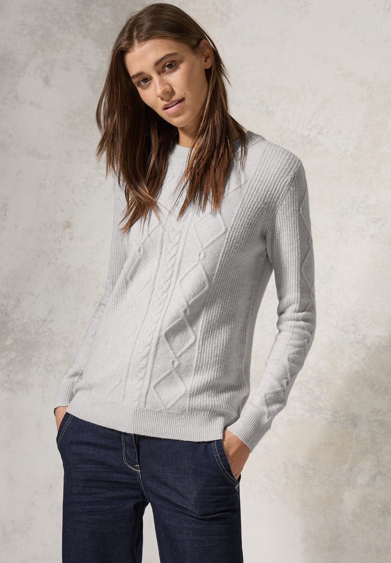 Maglione lavorato a maglia di un grigio chiaro con un motivo a rombi e dettagli a costine. Collo a giro e maniche lunghe, abbinato a jeans scuri.