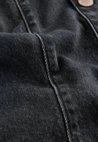 Tissu en denim foncé présentant un tissage texturé, avec des coutures claires contrastantes et un détail de bouton en cuivre visible le long du bord.