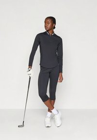 Μαύρο αθλητικό τοπ με φερμουάρ στον γιακά και μακριά μανίκια, σε συνδυασμό με εφαρμοστά καπρί κολάν. Λευκά αθλητικά παπούτσια και γκολφ κλαμπ ορατά.