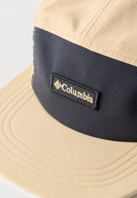 Μπεζ και σκούρο μπλε καπέλο Columbia με ορθογώνιο μπαλώμα κεντημένου λογότυπου στο μπροστινό πάνελ, που δείχνει λεπτομερή ραφή στο γείσο και τα πάνελ.
