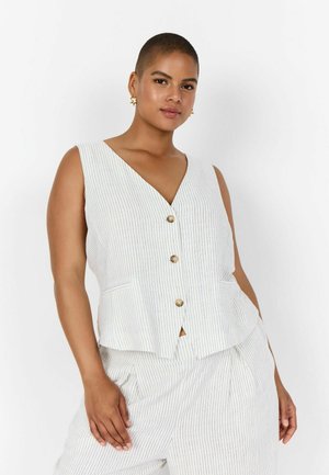 Frau mit kurzen Haaren, die eine weiße, ärmellose Weste mit Nadelstreifen und Knöpfen sowie passende Hosen trägt und mit einer Hand in der Tasche steht.