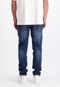 Man in blauwe spijkerbroek, witte sneakers en een wit T-shirt staat met zijn rug naar ons gekeerd tegen een witte achtergrond.