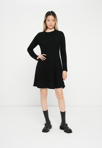 Vestido negro de manga larga con corsé ajustado y falda acampanada. Tejido suave con una textura lisa, hasta la rodilla, combinado con zapatos negros y calcetines grises.