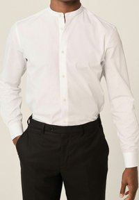 Uomo che indossa una camicia bianca con colletto alla mandarina, infilata in pantaloni eleganti neri, con le mani nelle tasche.