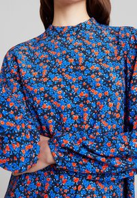 Femme les bras croisés portant une robe  à manches longues et col montant avec un motif dense de petites fleurs bleues et orange sur fond noir.