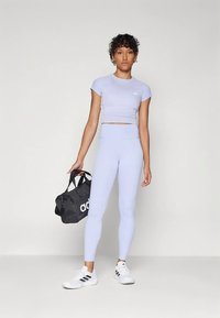 Nainen vaalean purppurassa urheilullisessa crop topissa ja legginsseissä, pitäen mustaa Adidas-urheilukassia, jalassa valkoiset Adidas-tennarit, seisoo tasaista taustaa vasten.