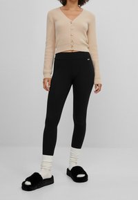 Beige pletený cardigan s knoflíky, černé legíny a černé huňaté šlapky v kombinaci s bílými pruhovanými ponožkami. Jednoduchý a přiléhavý design.