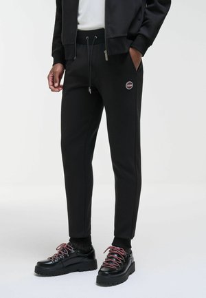Muž má na sebe čierne jogger nohavice s logom Colmar, čiernu bundu, bielu košeľu, čierne ponožky a čierne kožené topánky s červenými šnúrkami.