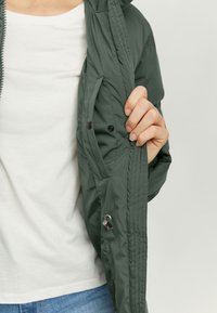 Green, lightweight jacket with a hood, featuring a zipper and snap buttons. Visible inner pocket with stitching detail. Soft texture.