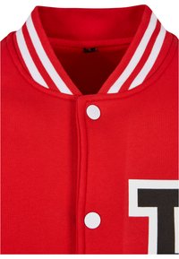 Rød varsity-jakke med hvite striper på kragen, med snapsknapper og en fremtredende svart og hvit bokstavlapp foran.