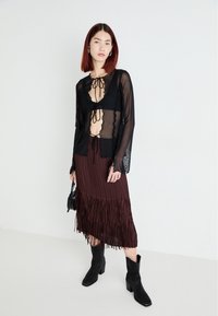 Czarny przezroczysty top z długim rękawem i wiązaniem z przodu, w połączeniu z brązową spódnicą midi z frędzlami. Modelka nosi czarne botki za kostkę i trzyma małą czarną torbę.