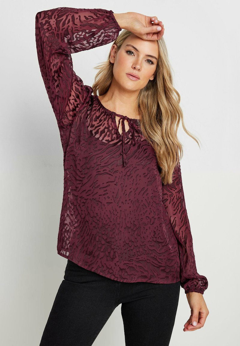 Blusa trasparente bordeaux con stampa animale texture, colletto con fiocco e polsini elastici. Maniche lunghe e vestibilità rilassata. Indossati pantaloni neri.