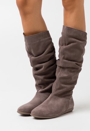 Zachte bruine suède slouchy kniehoge laarzen met vlakke zolen, gedragen op blote benen, tegen een effen witte achtergrond.