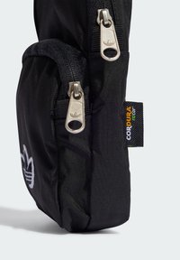 Svart tygväska med silverdragkedjor, texturerat material och en logopatch. Sidetiketten anger att den är tillverkad av hållbart Cordura-tyg.