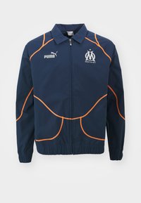 Mornarica jakna z oranžnim šivom in poudarki, ki ima visok ovratnik, zadrgo in logotipe na prsih. Gladka tekstura.