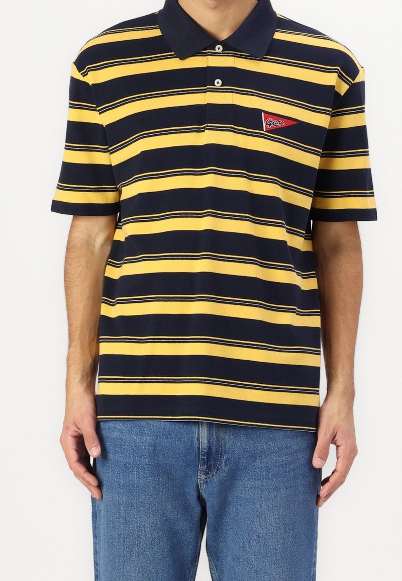 Homme portant un polo à manches courtes rayé jaune et bleu marine avec un écusson rouge et un jean bleu, debout sur fond blanc.