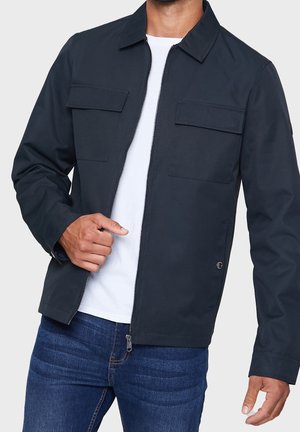 Homme portant une veste zippée bleu marine par-dessus un t-shirt blanc et un jean bleu, les mains ajustant la fermeture éclair de la veste.