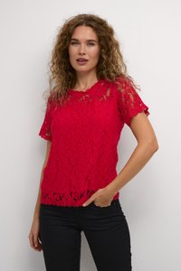 Top en dentelle rouge avec des manches courtes, des bords festonnés et des motifs floraux complexes. Porté sur un pantalon noir, mettant en valeur une coupe ajustée.