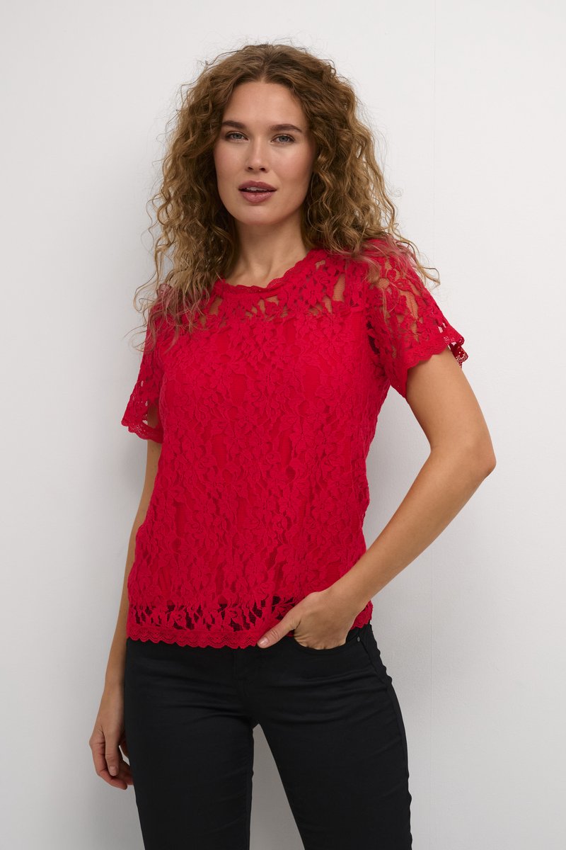 Top en dentelle rouge avec des manches courtes, des bords festonnés et des motifs floraux complexes. Porté sur un pantalon noir, mettant en valeur une coupe ajustée.