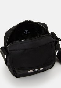 Mala crossbody preta com um tecido texturizado, fecho de zíper e uma etiqueta interna. Inclui uma alça ajustável e um bolso com aba na parte da frente.