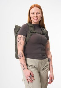 Olive green backpack with adjustable straps and side buckles, made of durable material. Person wears a fitted brown shirt and light-colored pants.