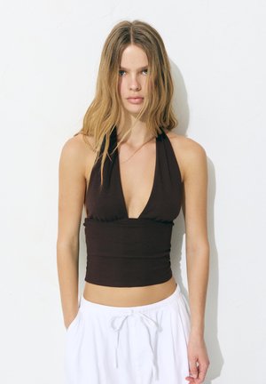 Junge Frau mit langen blonden Haaren, die einen schwarzen Haltertop und weiße Hose mit Kordelzug trägt, steht vor einem weißen Hintergrund.