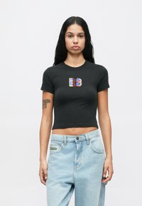 Γυναίκα με μακριά μαύρα μαλλιά που φοράει ένα μαύρο κοντό μπλουζάκι με το νούμερο "23" τυπωμένο και ανοιχτό μπλε τζιν, στέκεται μπροστά σε λευκό φόντο.