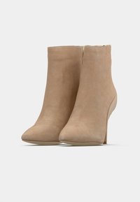 Cizme bej din piele întoarsă, de gleznă, cu vârf ascuțit și toc înalt subțire. Textură netedă, design minimalist și fermoar auriu accentuat în spate.