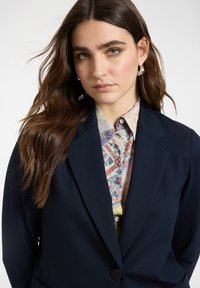 Joven con cabello largo y castaño que lleva pendientes de aro plateados, una chaqueta azul marino y una camisa abotonada con estampado floral multicolor.