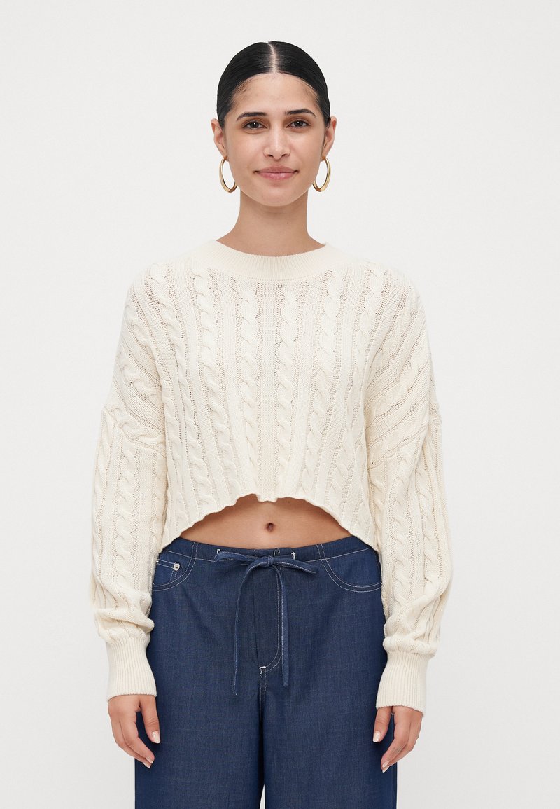 Woman with dark hair in a low bun wearing gold hoop earrings, a cream cable-knit cropped sweater, and high-waisted navy drawstring pants.